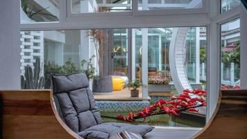 Un intérieur moderne avec une fenêtre incurvée, un fauteuil rembourré de couleur grise et une vue sur un espace extérieur agrémenté de plantes et d’un élément décoratif aquatique.