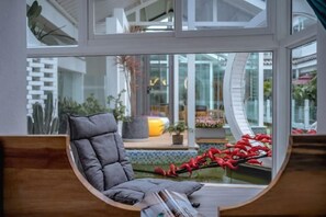 A modern interior with a curved window, a grey cushioned chair, and a view of an outdoor area with plants and a water feature. - Lushan Huadian Biyuan Theme Hotel (Weishan)
