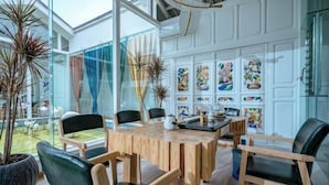 Un espace de salle à manger moderne, avec une table et des chaises en bois, entouré de murs en verre et offrant une vue sur un jardin.