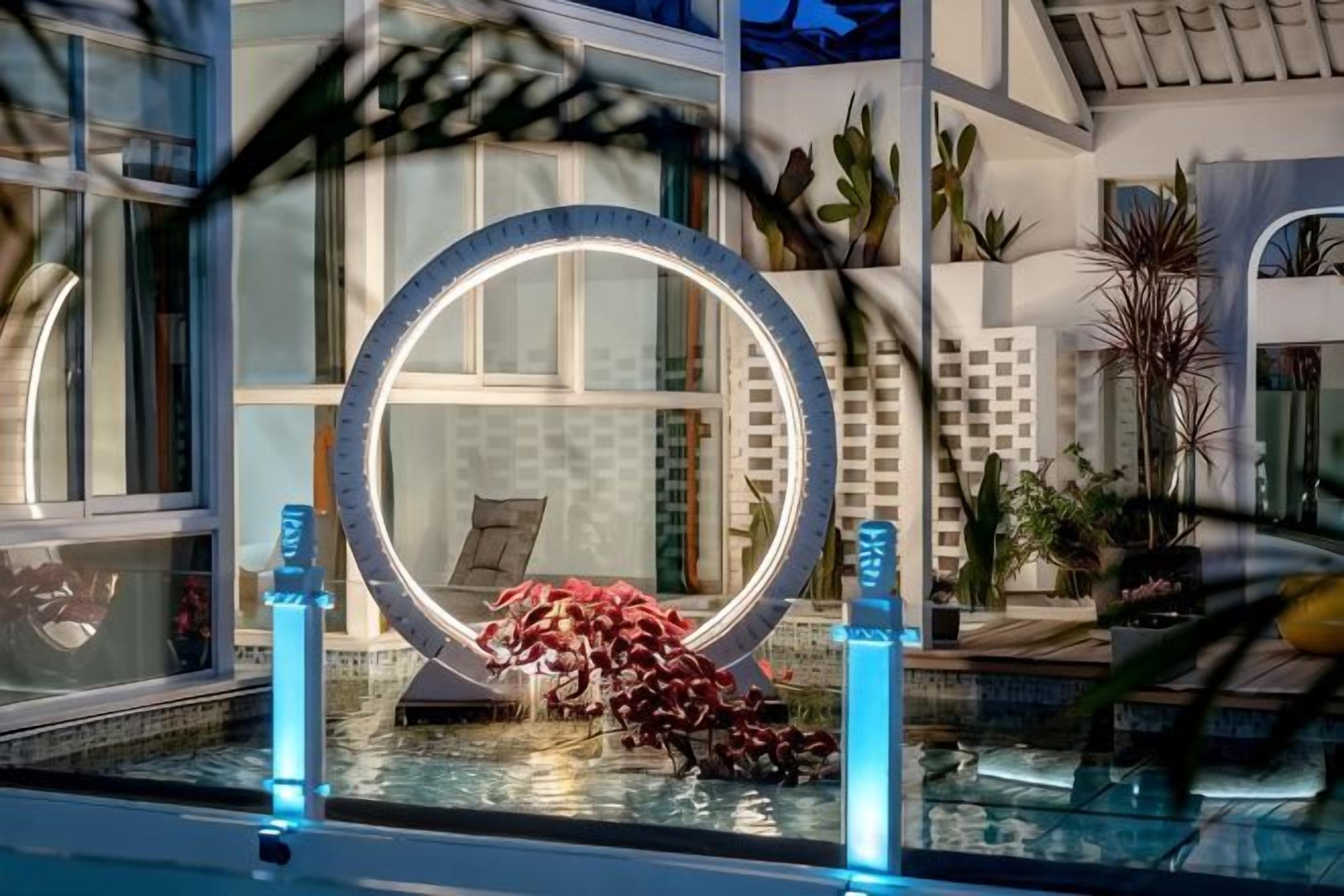 A pool area with a circular mirror, surrounded by plants and a tiled wall.