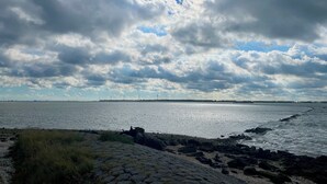Un paysage côtier avec une promenade pavée menant à l’eau, une côte rocheuse et une vue lointaine sur des éoliennes.
