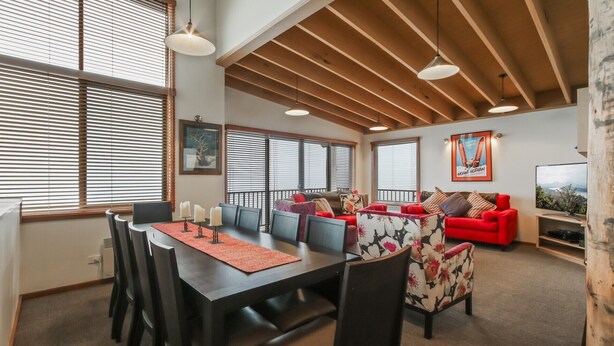 Una sala de estar moderna con comedor, vigas de madera en el techo y grandes ventanales con persianas.