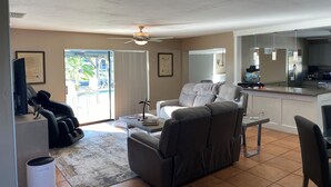 Ein Wohnzimmer mit Sofa, Couchtisch und Fernseher. Eine Glastür führt auf eine Terrasse mit Gartenmöbeln.