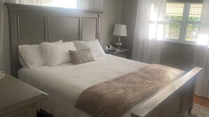 Ein ordentlich bezogenes Bett mit weißen Leinentüchern und einer braunen Tagesdecke, ein Nachttisch mit einer Lampe und ein Fenster mit Vorhängen.