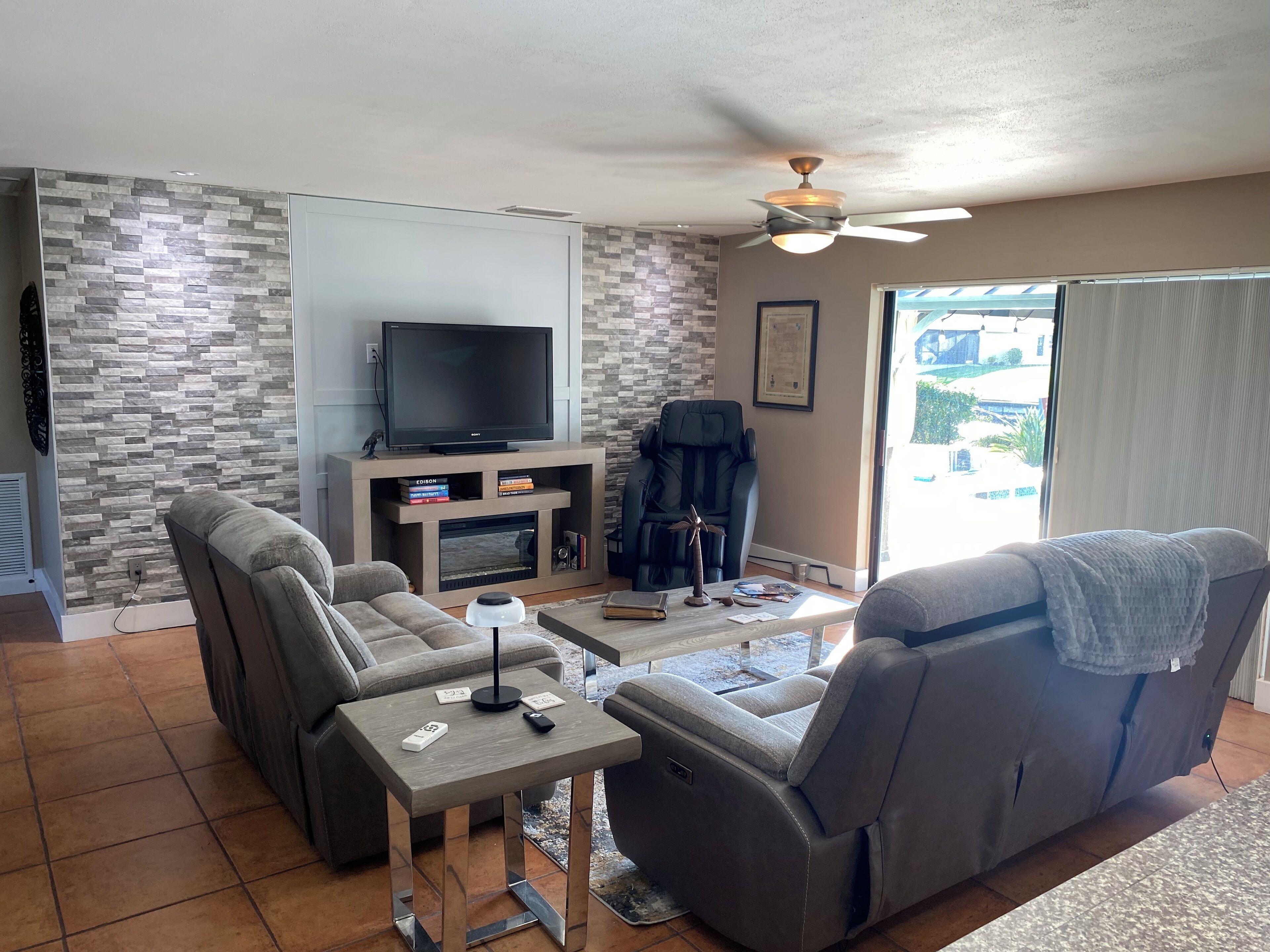 A living room with a flat-screen TV, a fireplace, a sofa, a recliner, and a ceiling fan.