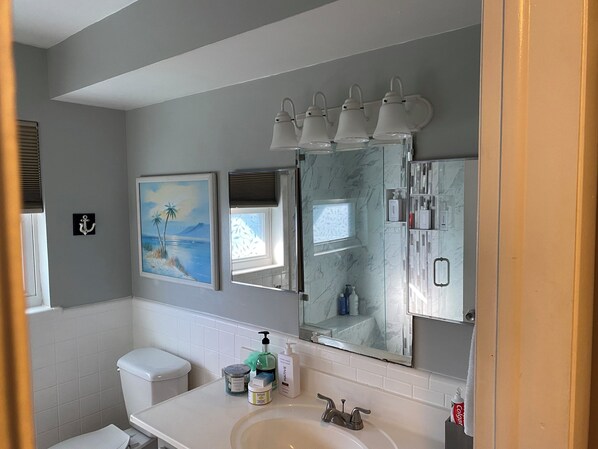 A bathroom with a toilet, sink, and three mirrors. There are framed pictures on the wall, a window, and a shower with a glass door.