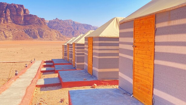 A row of tents with wooden doors and striped walls, set against a desert landscape with mountains in the background.