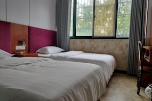 A hotel room with two beds, a wooden headboard, a window with curtains, and a small table with a lamp.