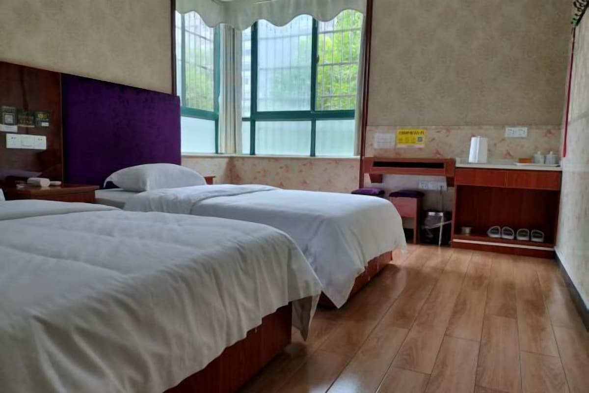 A room with two beds, a wooden floor, a window with curtains, a desk with a chair, and a small trash bin.