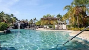 En klar pool med ett vattenfall, solstolar och palmer.