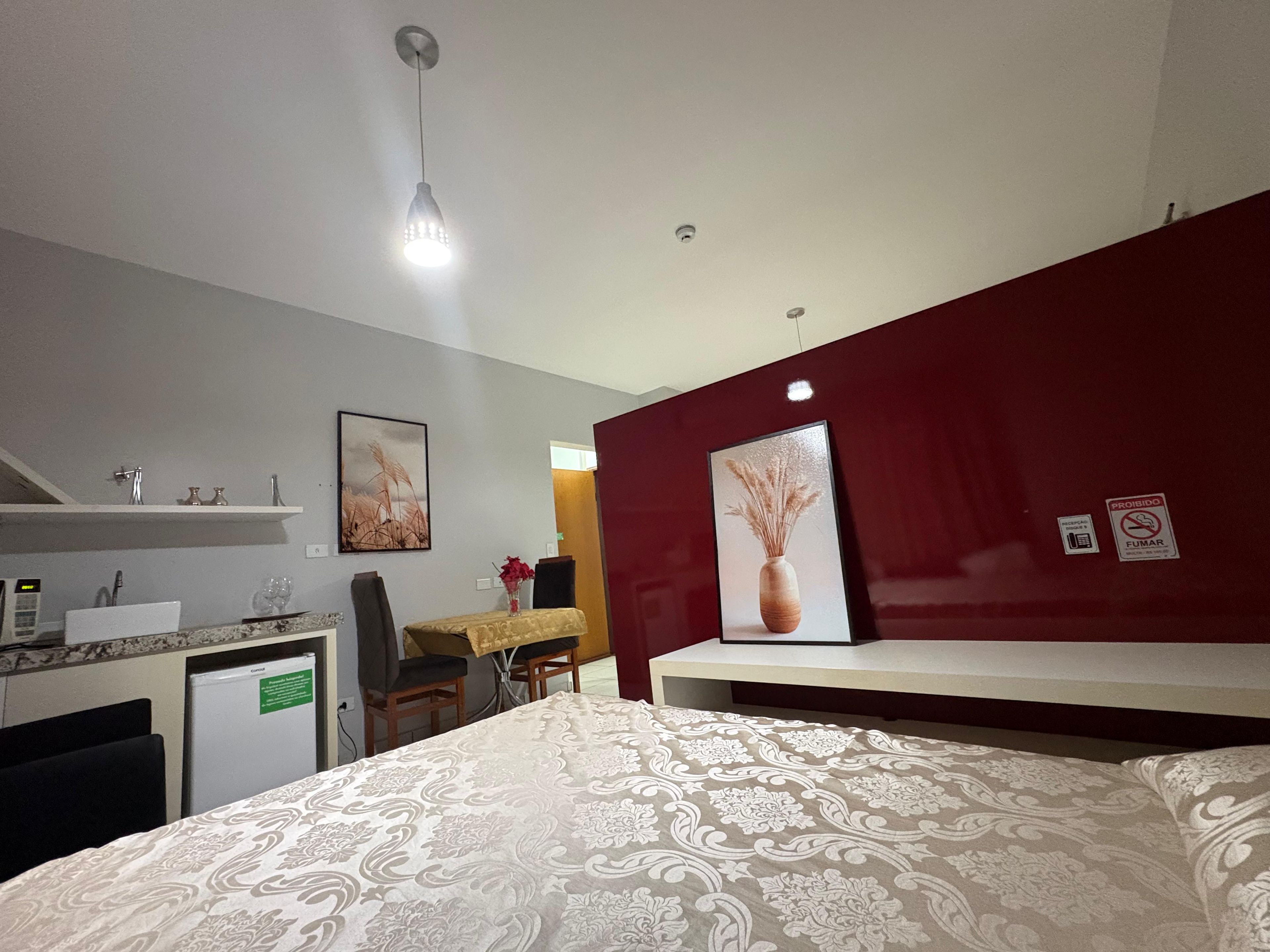 A hotel room with a bed, a desk, a chair, a small fridge, and a red accent wall.