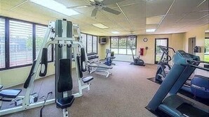 Een sportschool met diverse fitnessapparaten, een plafondventilator en grote ramen.