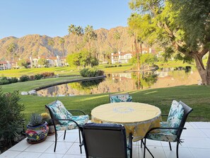 A patio with a round table and chairs, a potted plant, and a view of a lake with houses in the background.