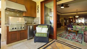 A kitchen with a stainless steel stove, wooden cabinets, and a wicker chair with cushions. - Beach club access, pool, Complex on A-bay, Private (Waikoloa Village)