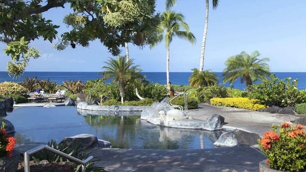 Ett poolområde på ett semesterhotell med en vattenfontän, palmer och utsikt över havet.