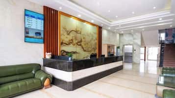 A hotel reception area with a reception desk, a green sofa, a decorative wall, and a staircase in the background.