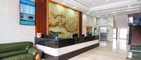 A hotel reception area with a reception desk, a green sofa, a decorative wall, and a staircase in the background.