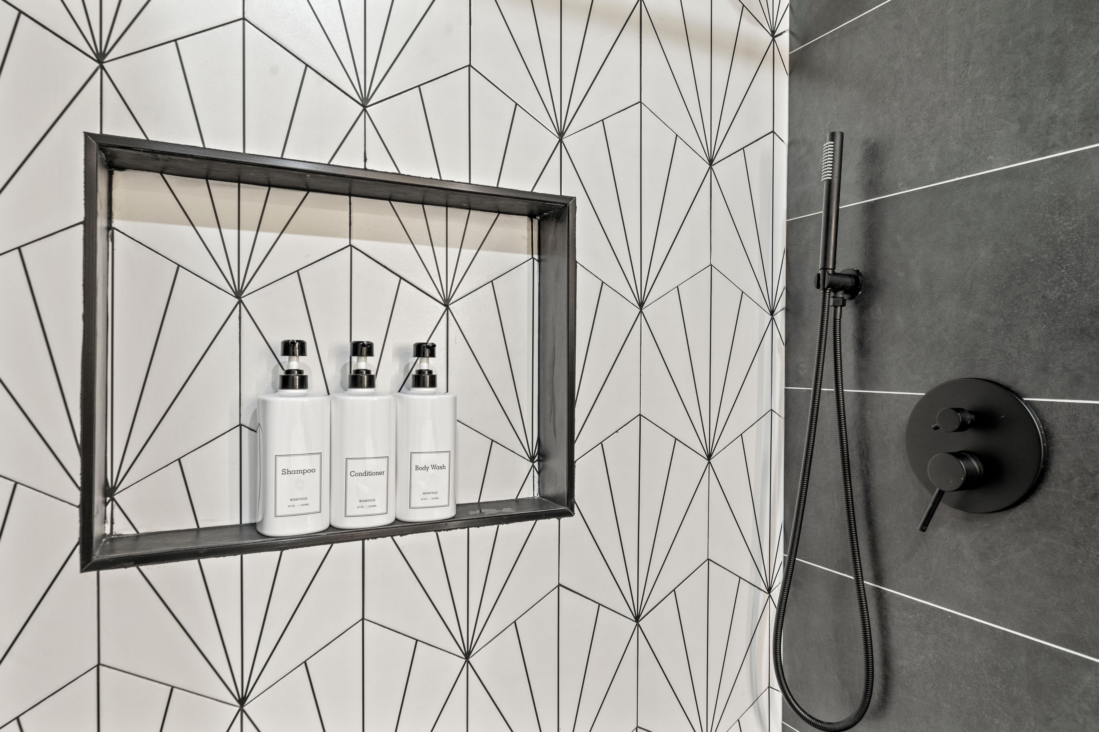 Ein Badezimmer mit Fliesenrückwand im geometrischen Design, einem Bilderrahmenregal mit drei Flaschen Shampoo, Conditioner und Duschgel sowie einer Handbrause.