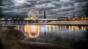 Eine Uferlandschaft mit Riesenrad und Leuchtturm, die sich in ruhigem Wasser unter einem bewölkten Himmel spiegelt.