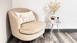 Ein beiger Sessel mit einem floralen Kissen, ein kleiner weißer Beistelltisch mit einer Vase getrockneter Blumen und eine Wasserflasche.