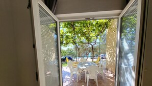 A sliding glass door leading to an outdoor patio with a table and chairs, surrounded by trees and a view of parked cars. - Apartments Radacic (Marina)