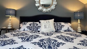A bedroom with a bed, two bedside tables, a wall-mounted mirror, and a pillow with the text "OUR NEST."