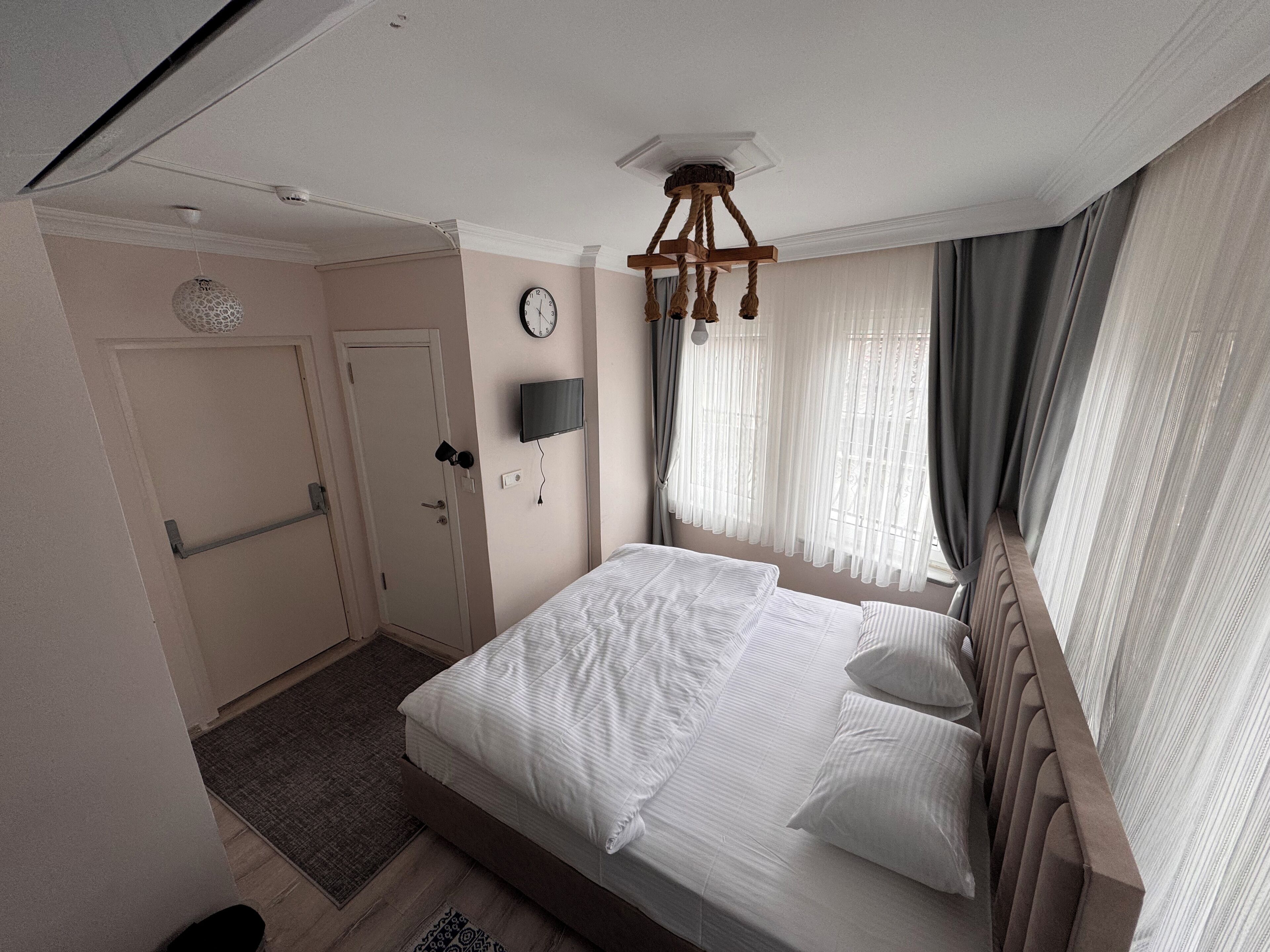 Habitación de hotel con una cama grande, televisión, reloj y ventana con cortinas.