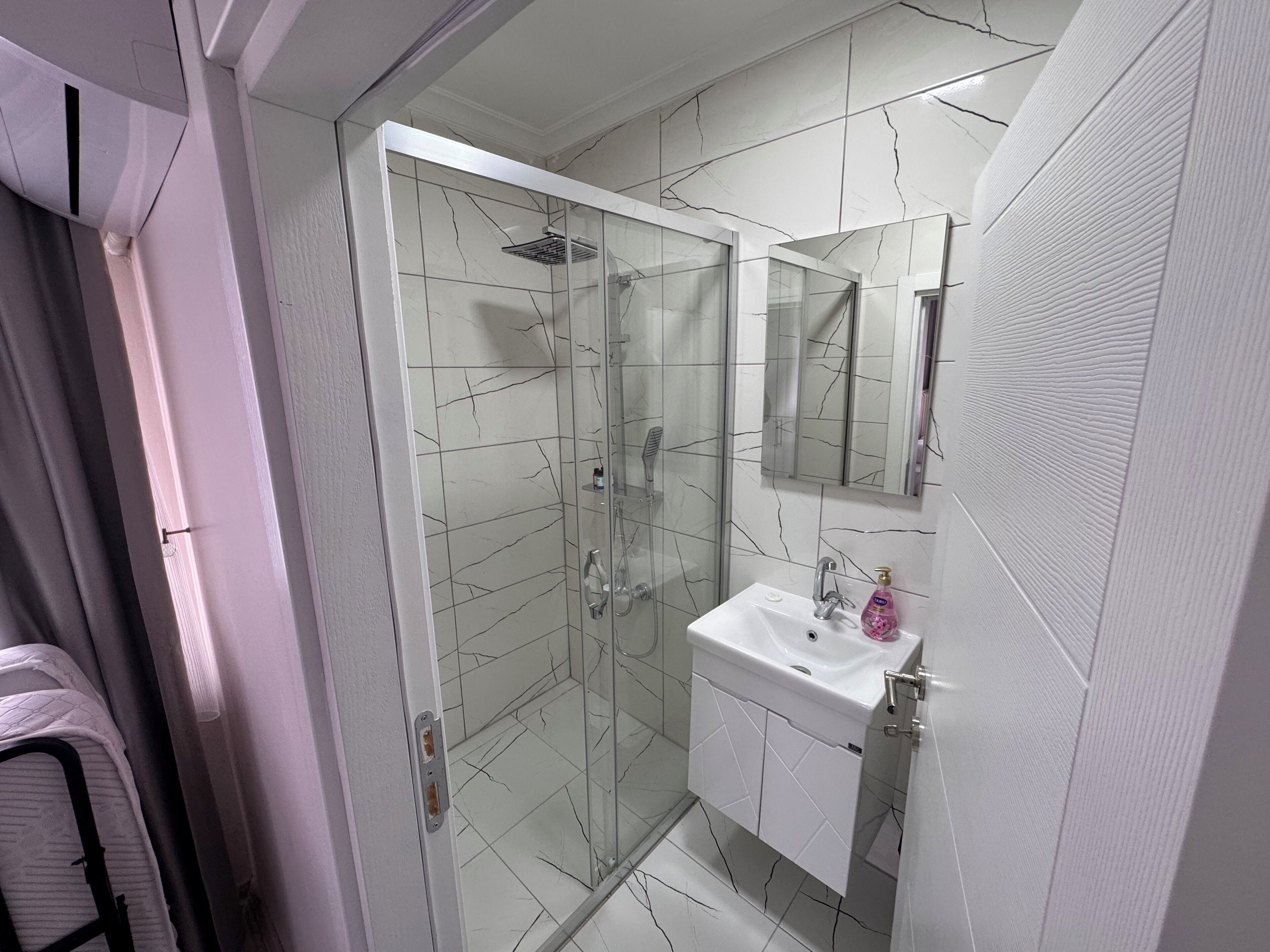 Un baño moderno con ducha de vidrio, un lavamanos blanco con espejo arriba y suelo de baldosas.