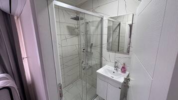 Un baño moderno con ducha de vidrio, un lavamanos blanco con espejo arriba y suelo de baldosas.