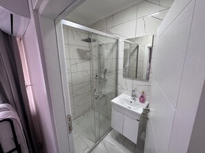 A modern bathroom with a glass-enclosed shower, a white sink with a mirror above it, and a tiled floor. - Villa Marina Mudanya (Mudanya)