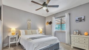 Ein Schlafzimmer mit Bett, Nachttischlampen, Deckenventilator, Kommode, Fenster mit Vorhängen und Wanddekorationen.