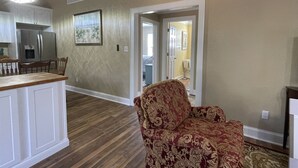 Een ruime woonkamer met een houten vloer, een fauteuil met een patroon erop, en een keuken met witte kasten en een roestvrijstalen koelkast.