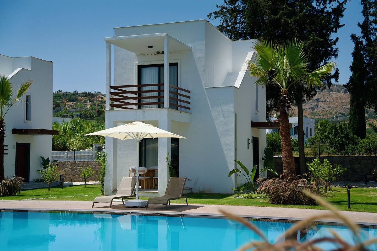 Ein moderner Poolbereich mit Liegestühlen und Sonnenschirm, umgeben von viel Grün und wolkenlosem, blauem Himmel.