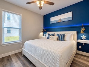 A bedroom with a bed, a ceiling fan, a window with blinds, a wall-mounted painting, and a nightstand with a lamp.