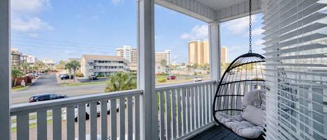 A porch with a hanging chair, a view of a street with cars, and buildings in the background.
