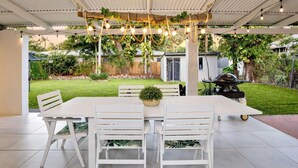 Um alpendre coberto com uma mesa e cadeiras brancas, luzes de cordão e vegetação.