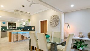 Uma zona de refeições moderna com mesa de vidro, cadeiras acolchoadas, uma peça decorativa na parede e uma cozinha ao fundo.