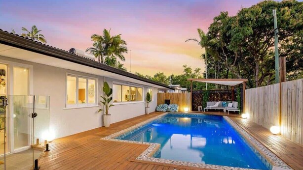 Piscina com deck de madeira, área de estar coberta e um prédio branco com janelas amplas.
