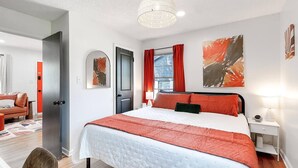 A modern bedroom with a large bed, a bedside table with a lamp, a door, and a window with red curtains.