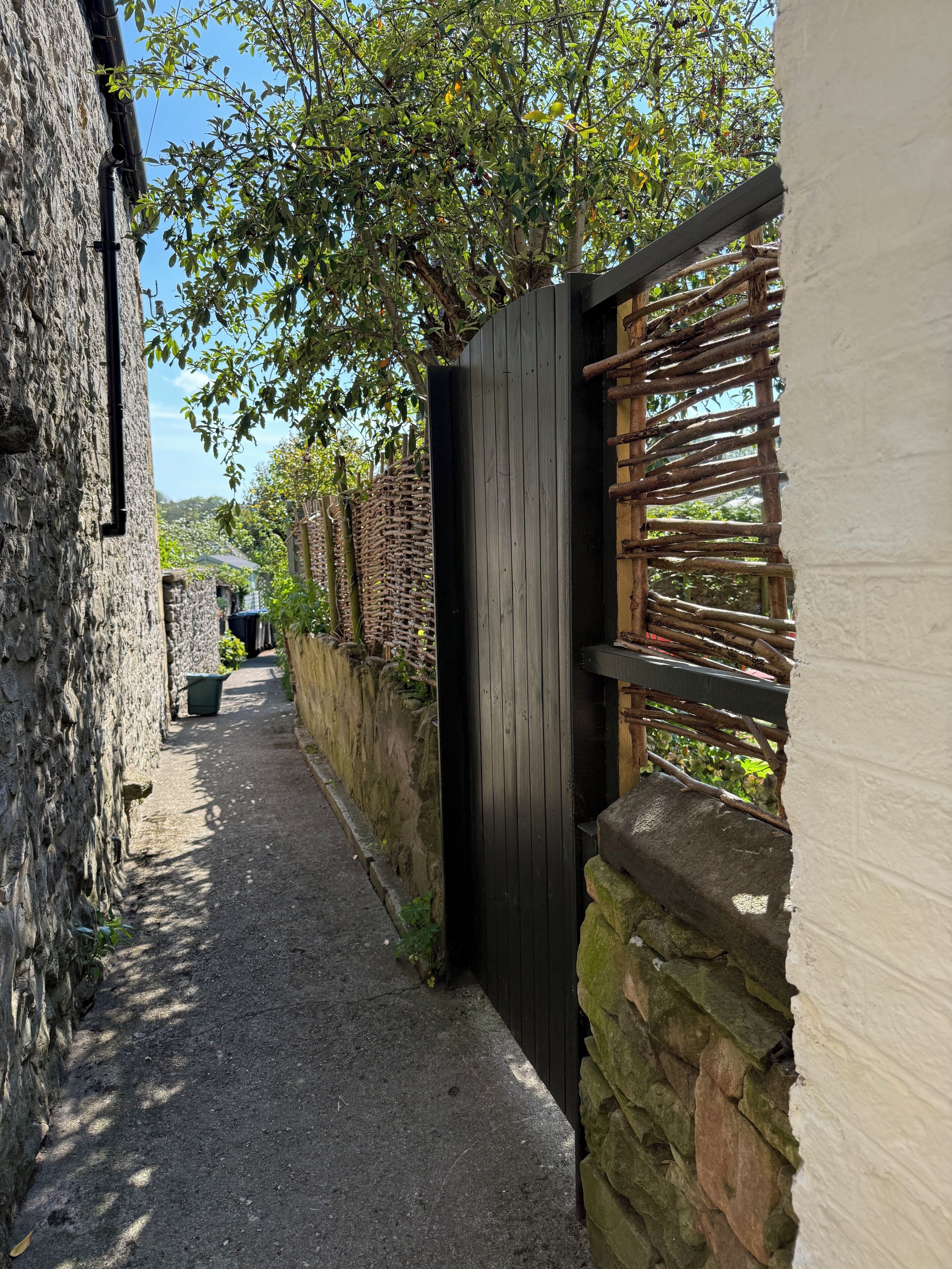 A narrow alleyway with stone walls, a wooden gate, and a tree providing shade.