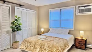 Un dormitorio con cama, dos mesitas de noche con lámparas, una planta en maceta, un ventilador de techo y una ventana con persianas.