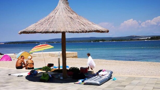 Een strand met een rieten parasol, een betegeld terras en mensen op het strand.