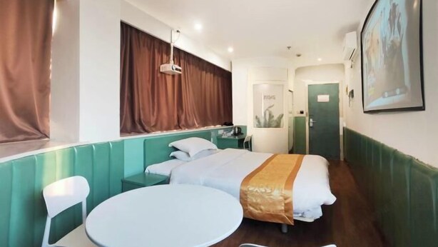 Hotelkamer met een bed, een rond tafel, een stoel, een raam met gordijnen, een deur en een aan de muur bevestigde projector.