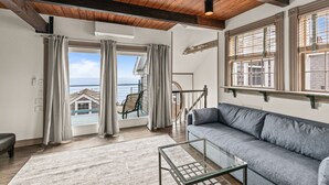 A living room with a view of the sea, a gray sofa, a glass coffee table, and a wooden ceiling.
