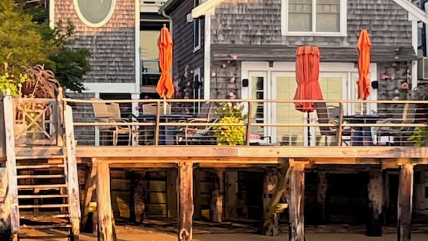 A waterfront property with a wooden dock, a stone building with a balcony, and orange umbrellas.