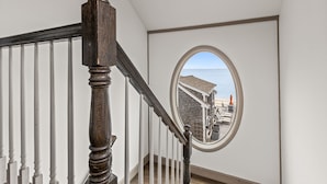 A staircase with a wooden handrail and white balusters, leading to a room with a large circular window offering a view of a deck and a building.
