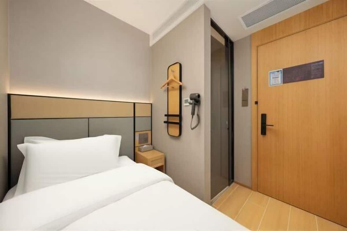 Quarto de hotel com cama, mesa de cabeceira, espelho, porta-toalhas, chuveiro e porta de madeira.