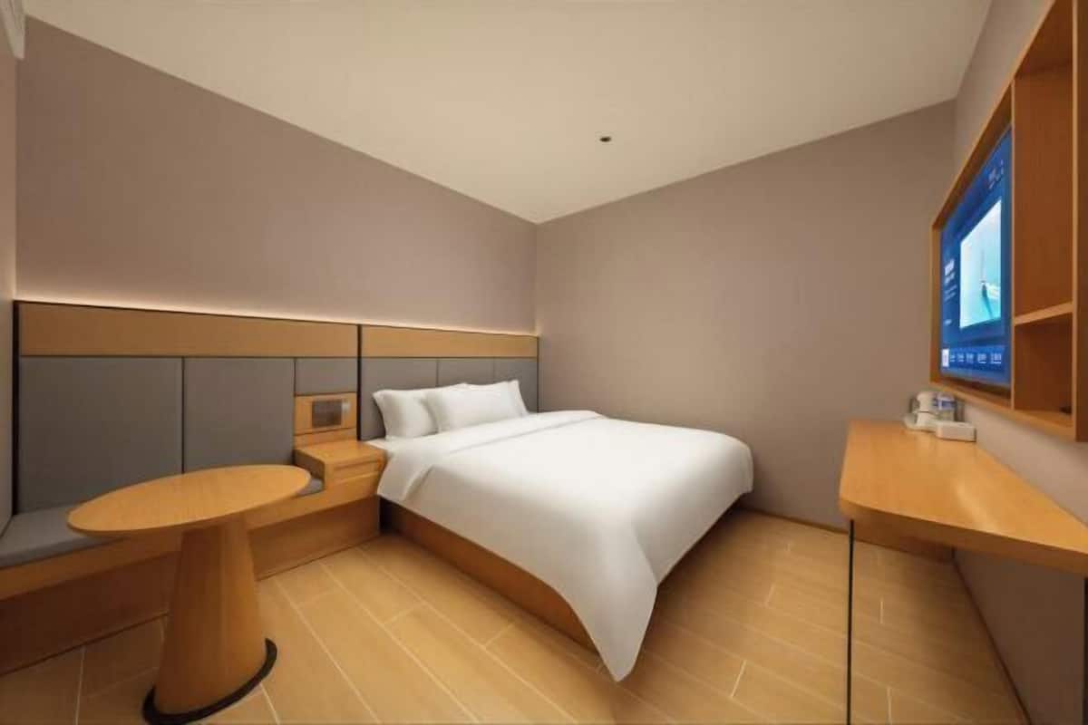 Quarto de hotel moderno com uma cama, uma mesinha redonda, um banco e uma televisão fixada na parede.