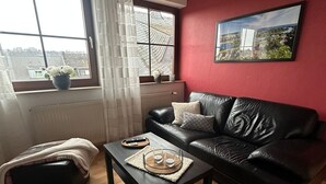 Un salon avec un canapé en cuir noir, une table basse avec des verres et un plateau, un radiateur, une fenêtre avec des rideaux et une vue sur le paysage urbain.