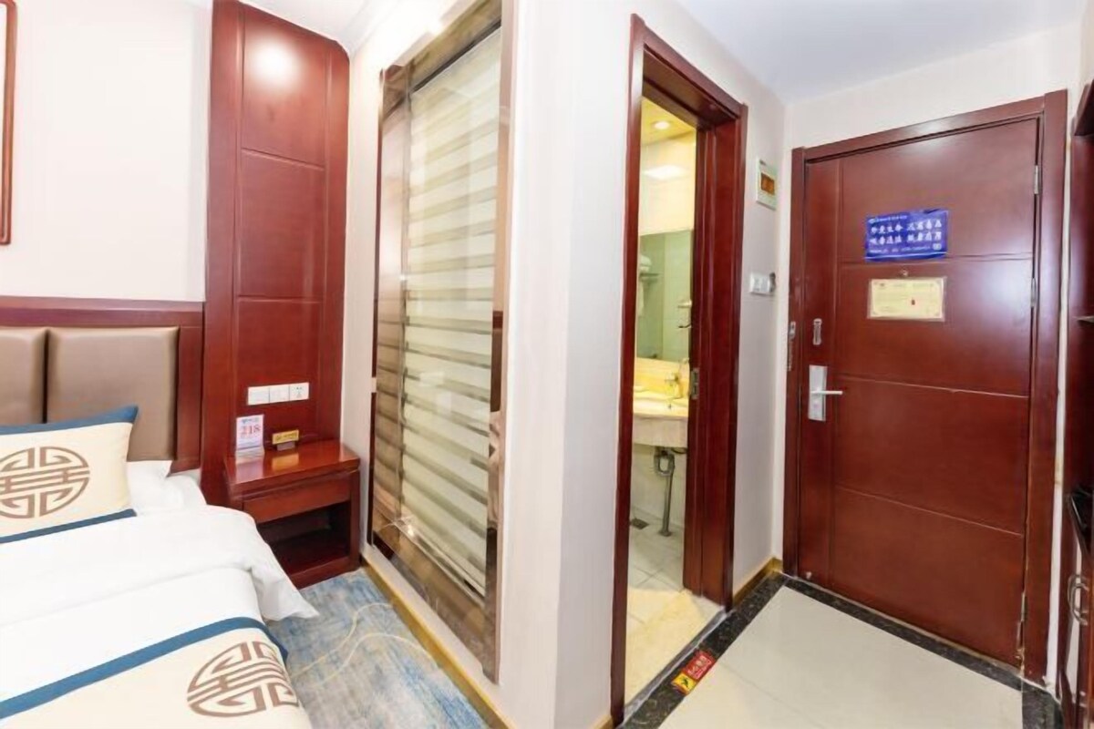 A hotel room with a bed, a wooden wardrobe, a bathroom visible through a glass door, and a wooden door with a sign.